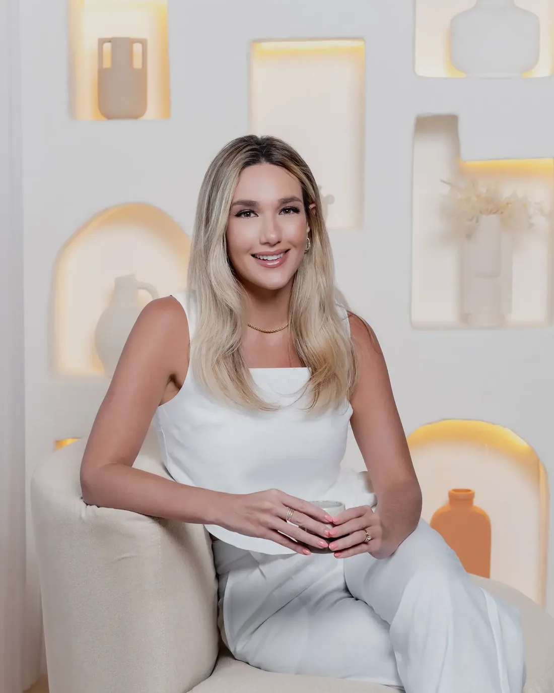 Dra. Vickie White, de branco, sentada numa poltrona clara diante de nichos iluminados, sorrindo.